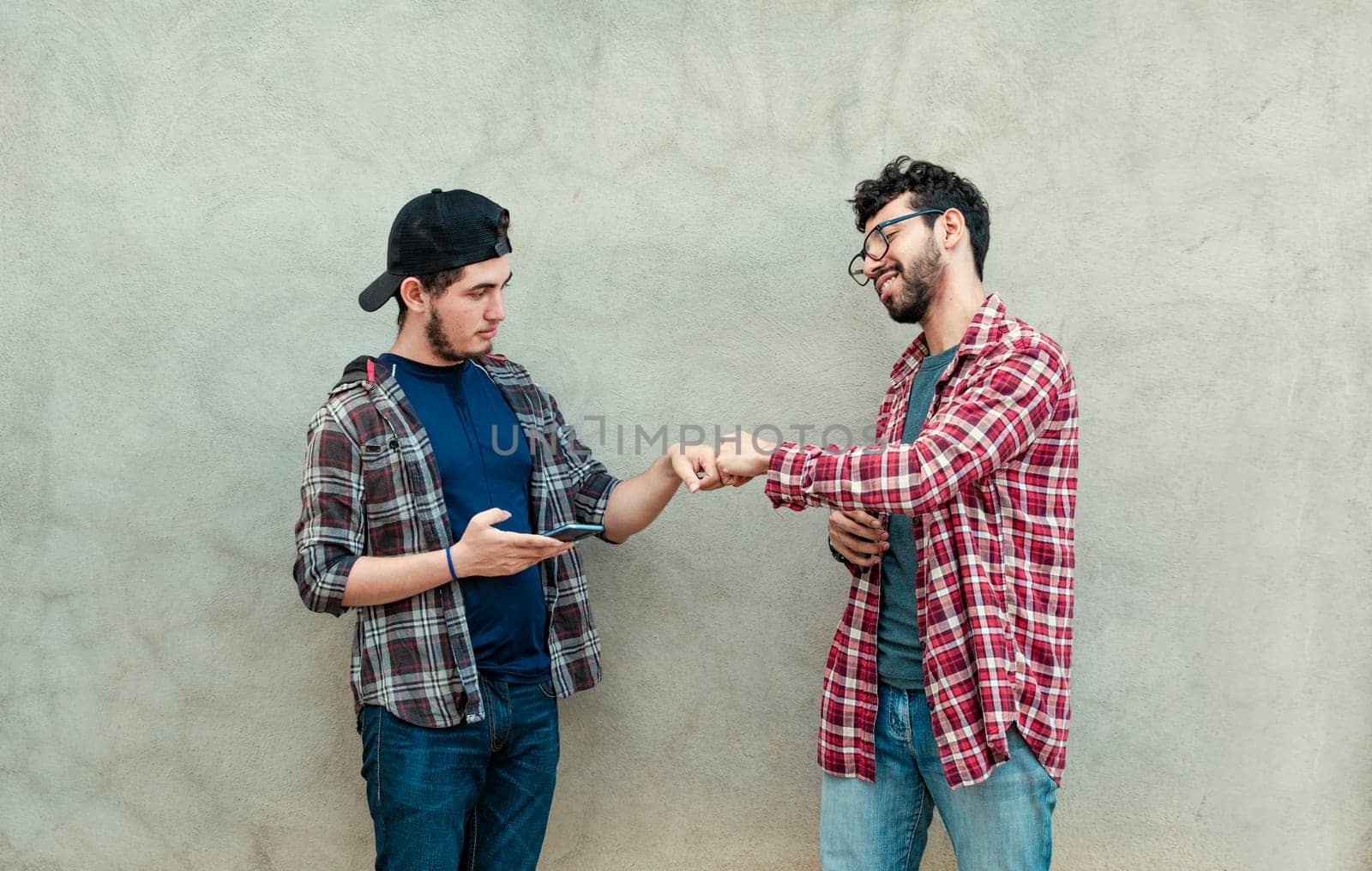 Two teenage friends greeting each other by fist bumping outdoors. Two smiling young guys bumping fists near a wall. Concept of two smiling friends bumping fists friendly