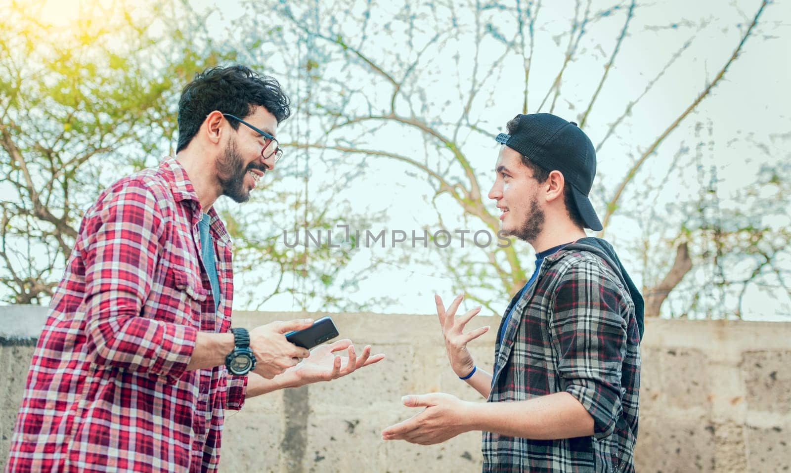 Two friends chatting while smiling and using cellphone. View of two teenagers talking face to face while using the cell phone, Side view of two young friends chatting outdoors