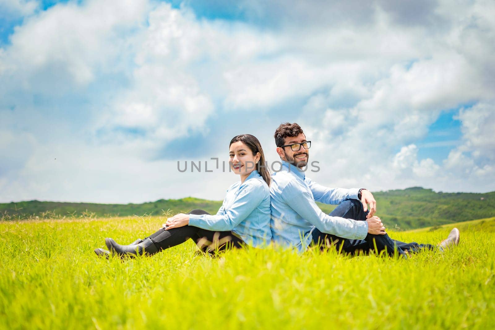 A wedding couple sitting with their backs to each other on the grass, Wedding couple in the field sitting with their backs to each other looking towards the camera