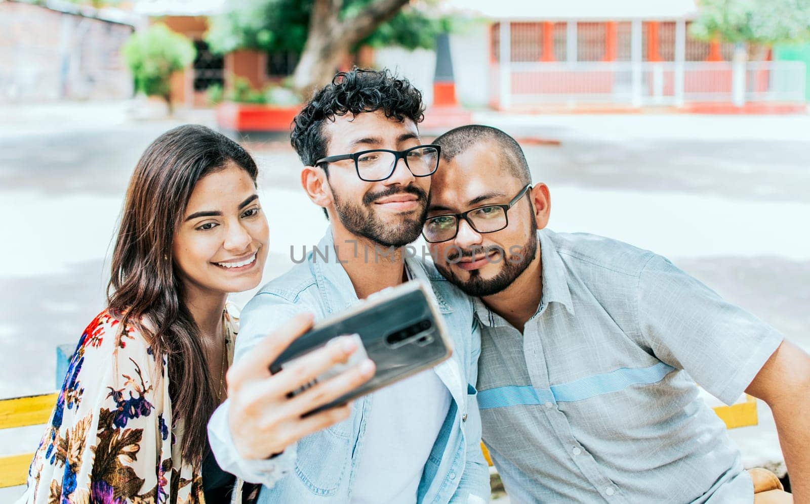 Three smiling friends sitting on a bench taking a selfie. Front view of three happy friends taking a selfie while sitting on a bench. Three friends taking a selfie on a park bench