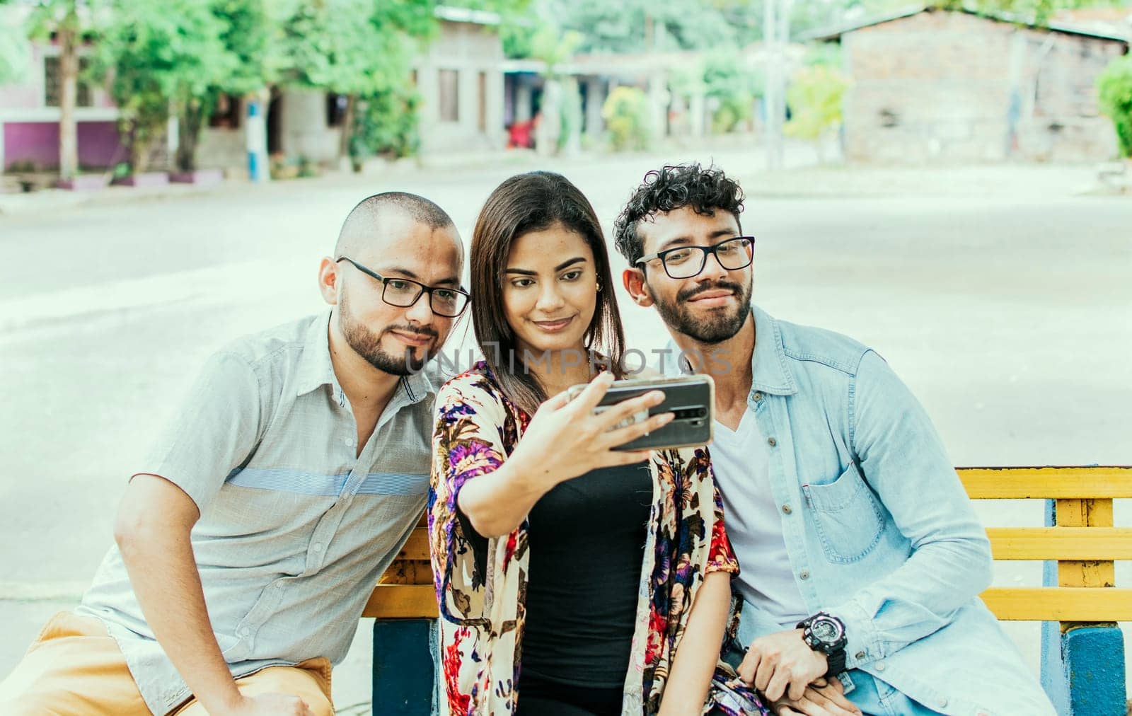 Three friends taking a selfie on a park bench, Three smiling friends sitting on a bench taking a selfie. Front view of three happy friends taking a selfie while sitting on a bench