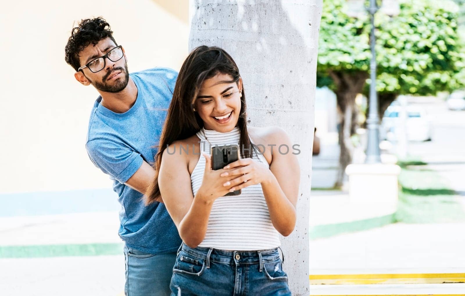 Jealous boyfriend spying on his girlfriend's cell phone in the park, Jealous man spying on his girlfriend texting on the cell phone. Suspicious man spying on his girlfriend with his cell phone