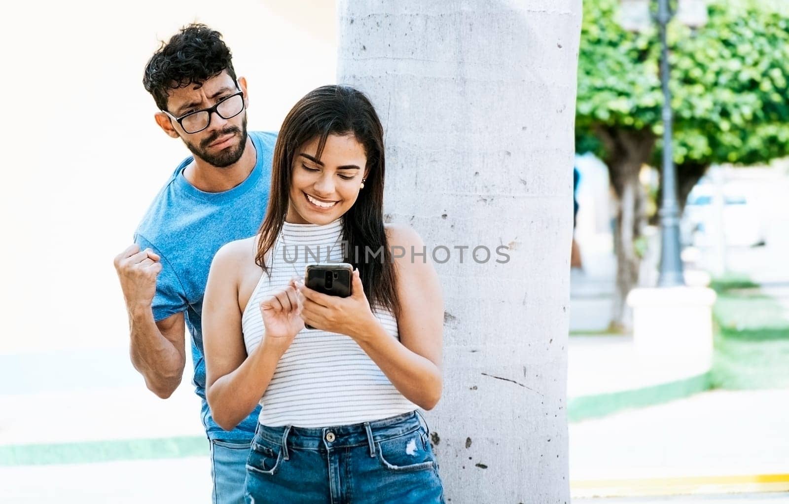 Jealous man spying on his girlfriend texting on the cell phone. Suspicious man spying on his girlfriend with his cell phone, Jealous boyfriend spying on his girlfriend's cell phone in the park