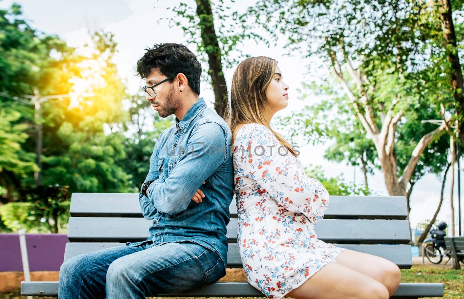 Upset young couple sitting back to back in a park, unhappy couple sitting back to back on bench. Concept of couple problems and crisis. Young couple arguing sitting back to back on a bench