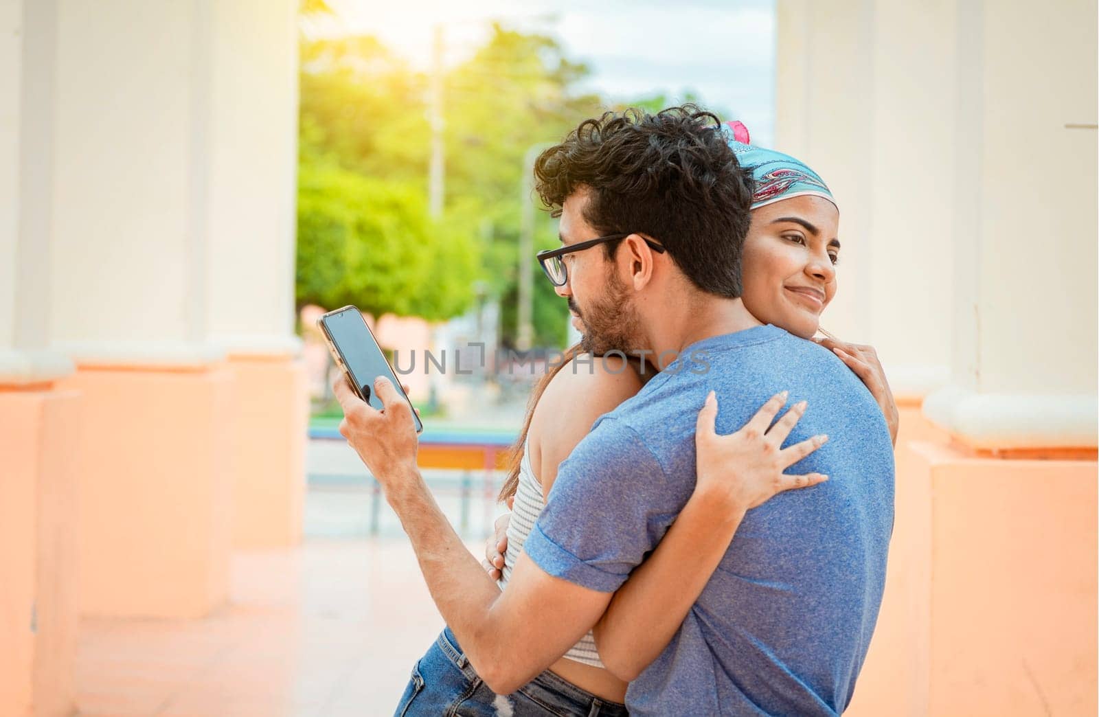 Unfaithful boyfriend hugging his girlfriend and looking at the phone. Unfaithful man looking cell phone while hugging his girlfriend outdoors, Concept of unfaithful man using cell phone
