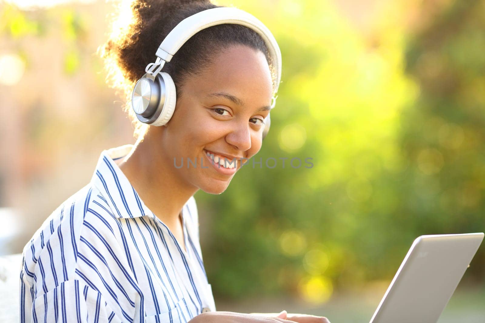 Happy black woman with laptop and headphone looking at you in a park