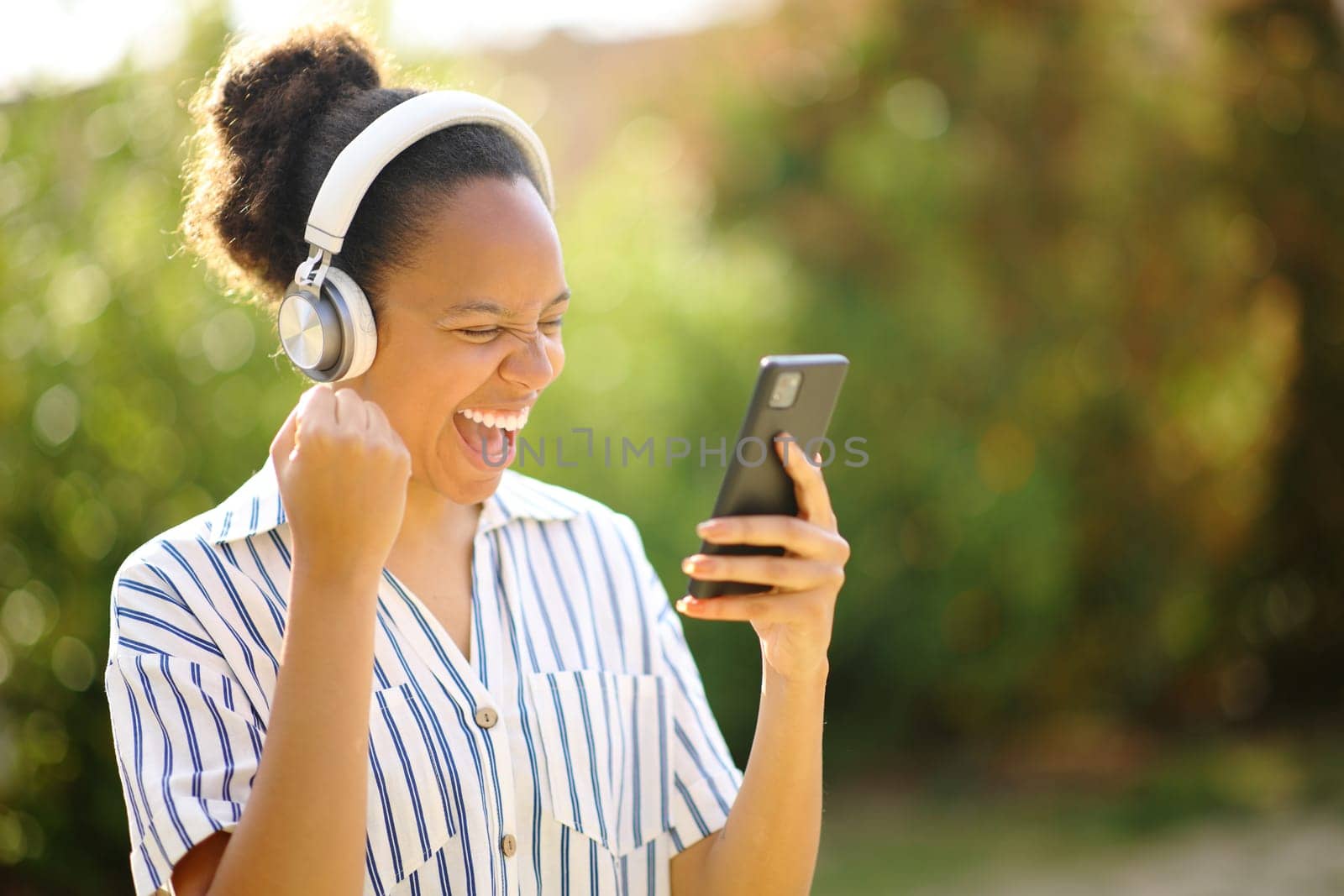 Excited black woman wearing headphone celebrating listening to music with phone in a park
