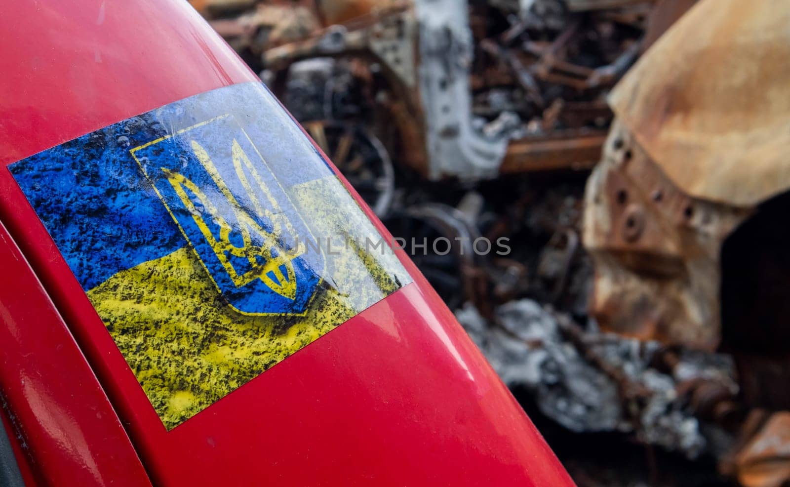 Sticker with Ukrainian flag and coat of arms. Close up yellow and blue sticker with the Ukrainian flag on a red car against the background of ruins and burnt cars. Ukraine, Kyiv - May 06, 2023