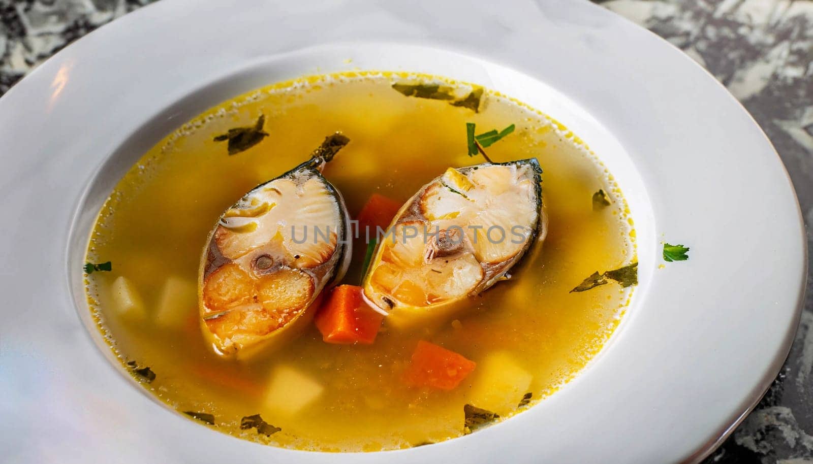 Fish soup Ukha from sturgeon. In a white, round, ceramic plate, there are two pieces of sturgeon fillet, chopped carrots and fish broth. Nearby is a small, white, ceramic bowl with finely chopped dill