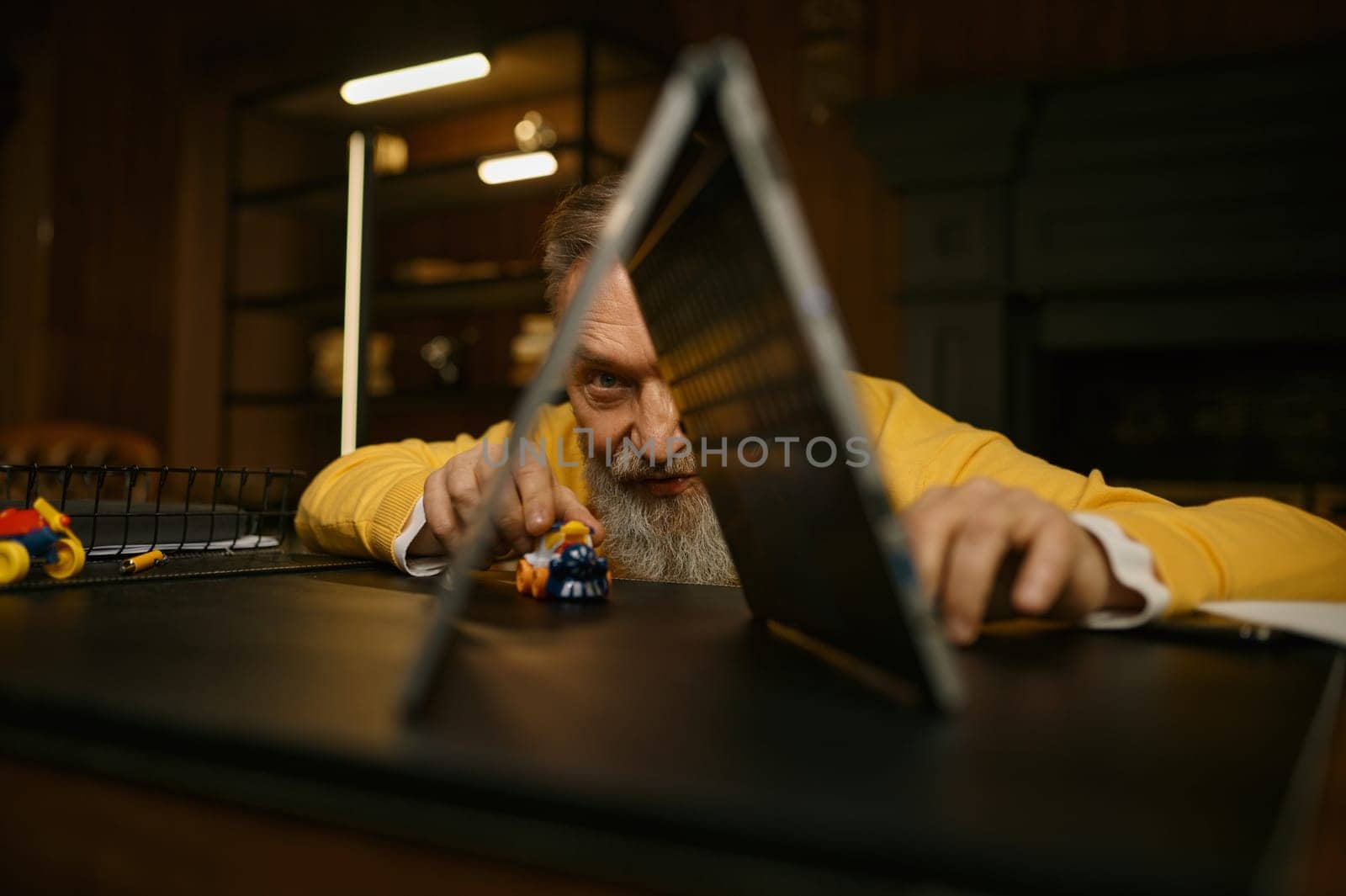 Senior man playing toys at workplace having fun after hard work in home office. Older employee accountant taking break time enjoying train race gaming