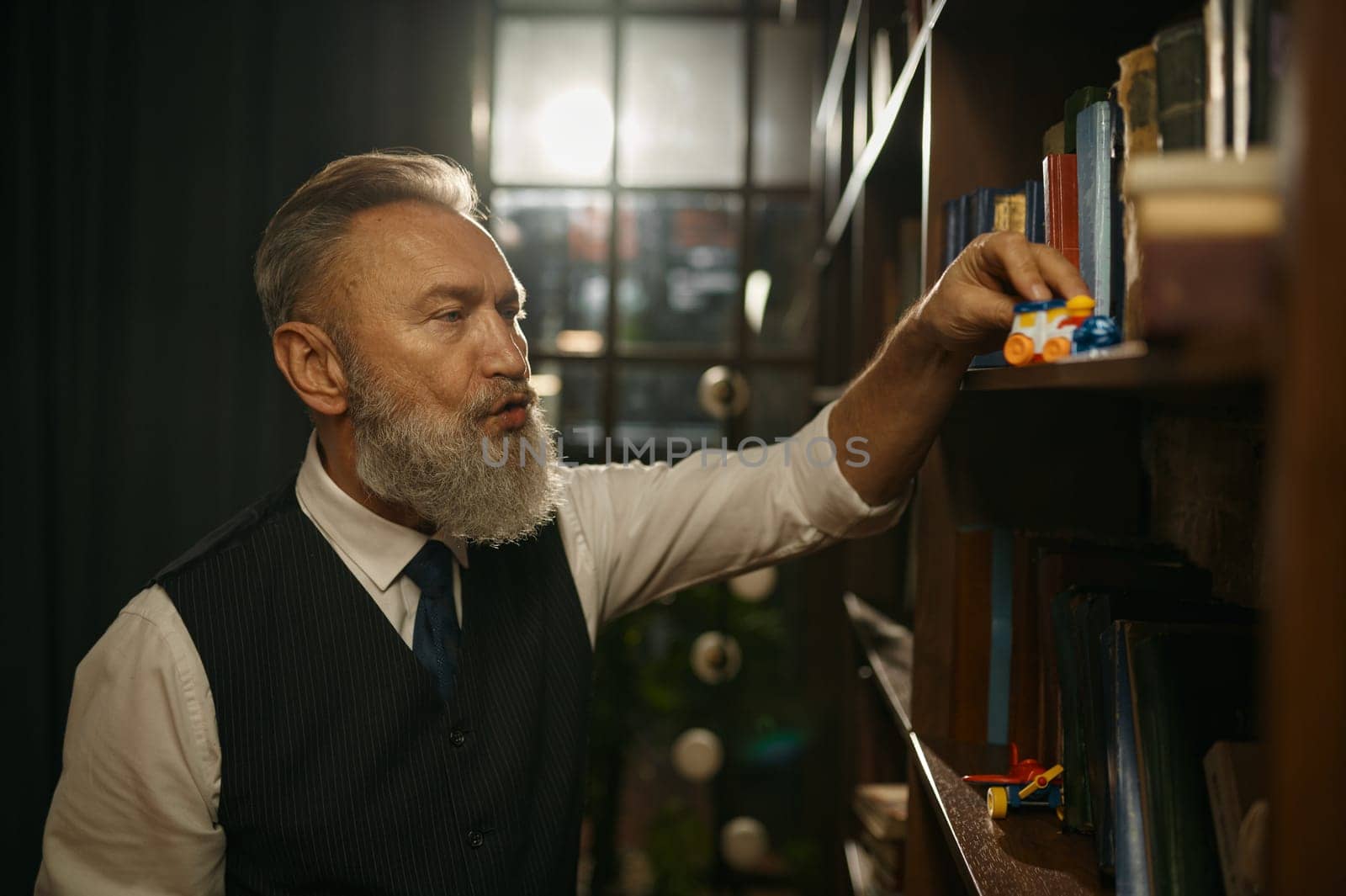 Senior businessman with old-fashioned outfit playing toy train on bookshelf in home or office library