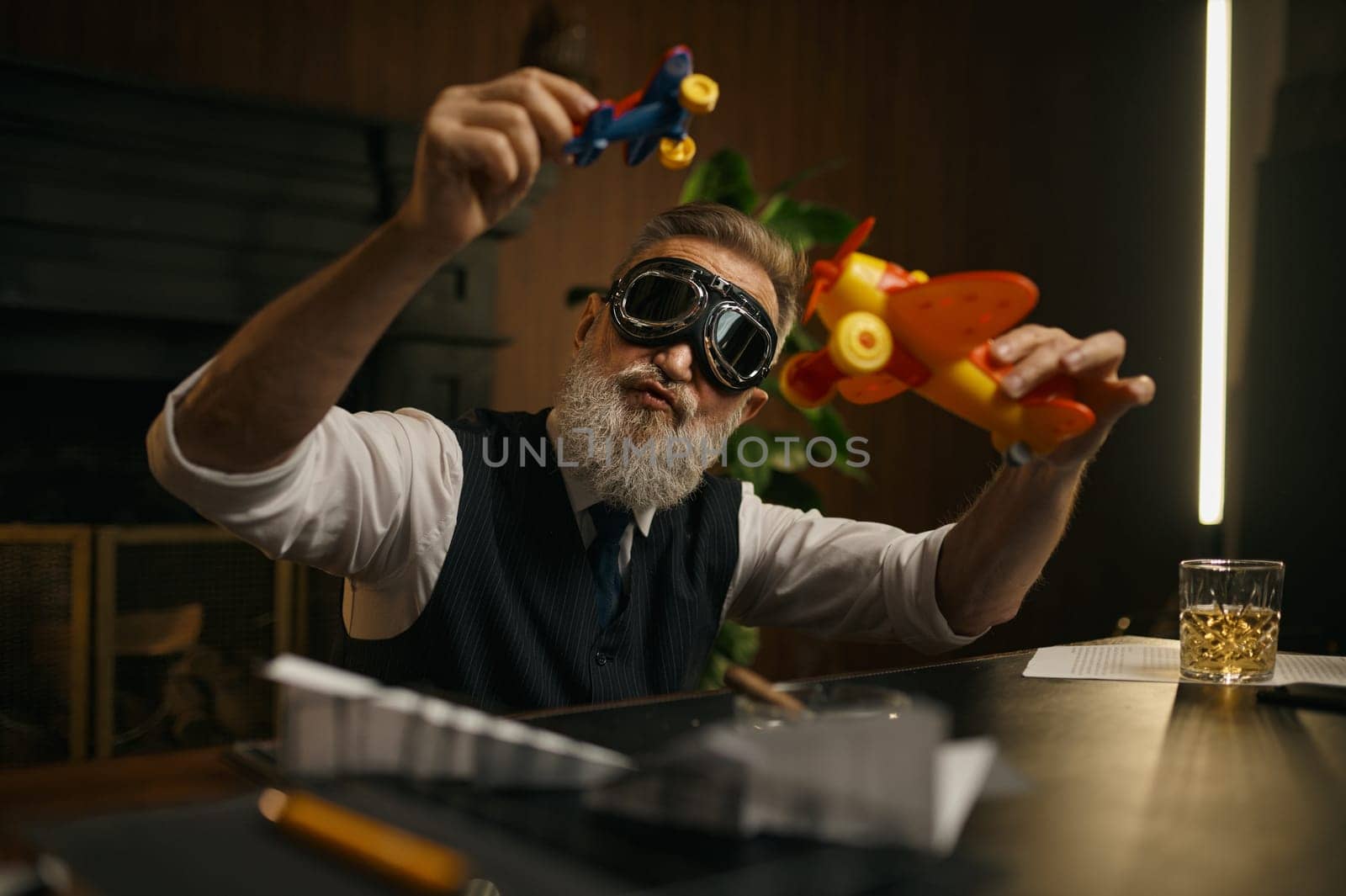 Playful senior boss playing toy plane at work table. Mature grey-haired businessman wearing elegant clothes and aircraft pilot goggles having fun and relax