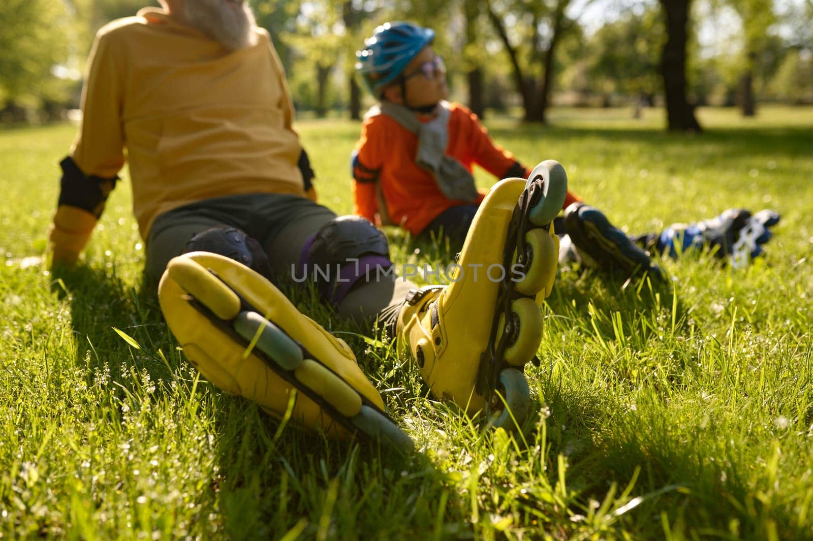 Happy man and boy rollerskaters rest on grass filed in park. Young and adult people having active leisure activity and fun sports time together