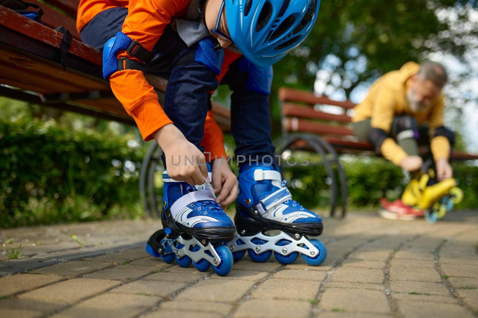 Selective focus of grandson and grandfather putting on roller skates. Mature man and boy preparing for racing in park playground