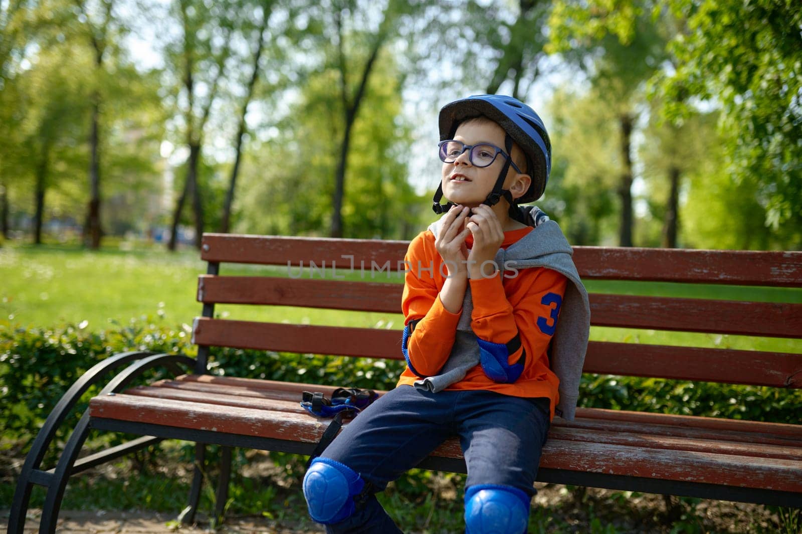 Little boy wearing protective helmet on head while sitting on bench in park. Preparation for safety extreme riding roller skates outdoors and active lifestyle concept