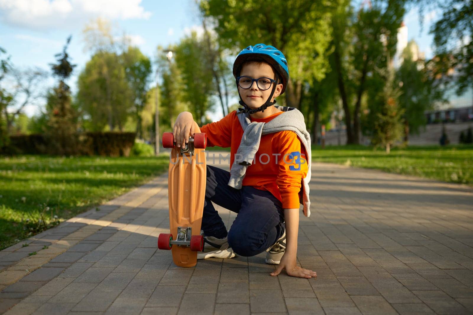 Portrait of smiling little schoolboy wearing protective helmet sitting on asphalt with skateboard. Young child athlete full length shot. Safety sport for children