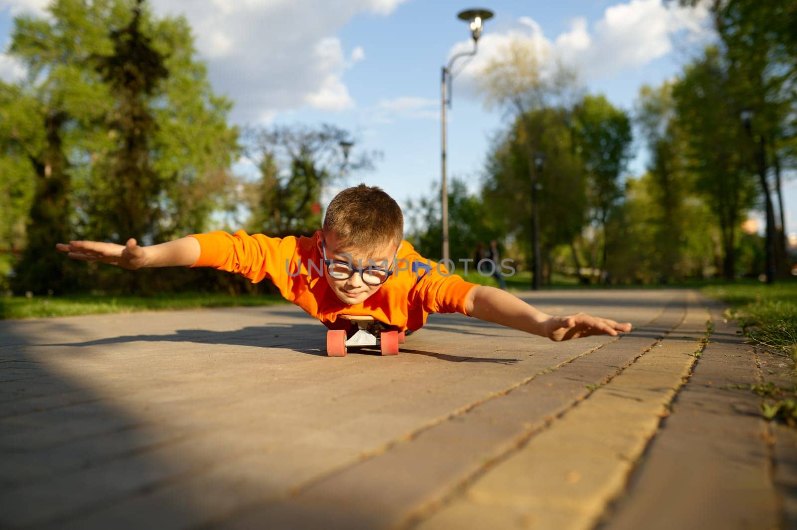 Boy lying on skateboard on his belly and riding like aviator pretending to fly in sunny park. Happy youngster enjoying activity lifestyle and extreme sport hobby