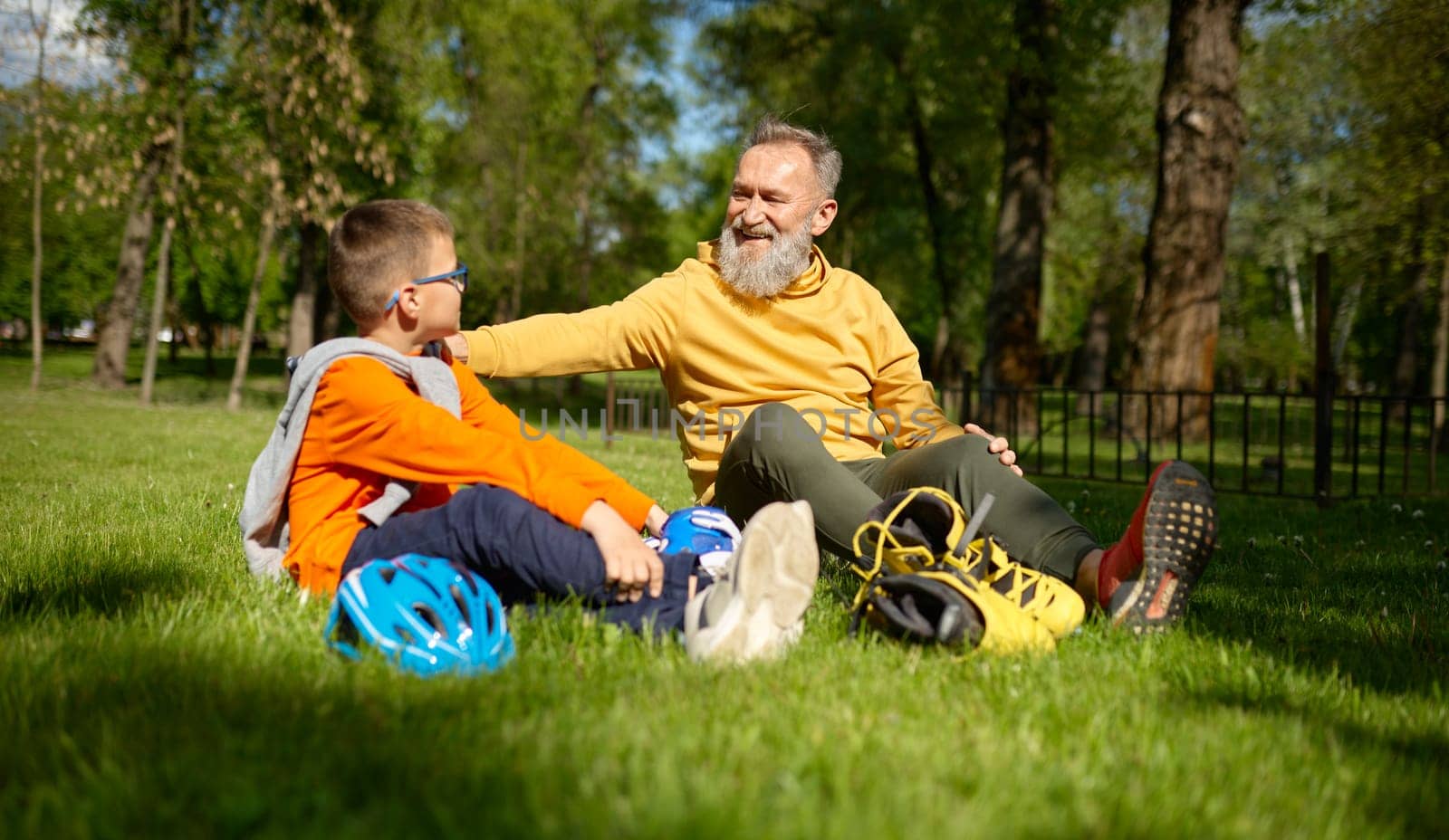 Positive smiling grandpa and grandson rest on green grass meadow in park. Granny supporting and praising grandchild for good riding roller-skates. Happy family recreation time on weekend