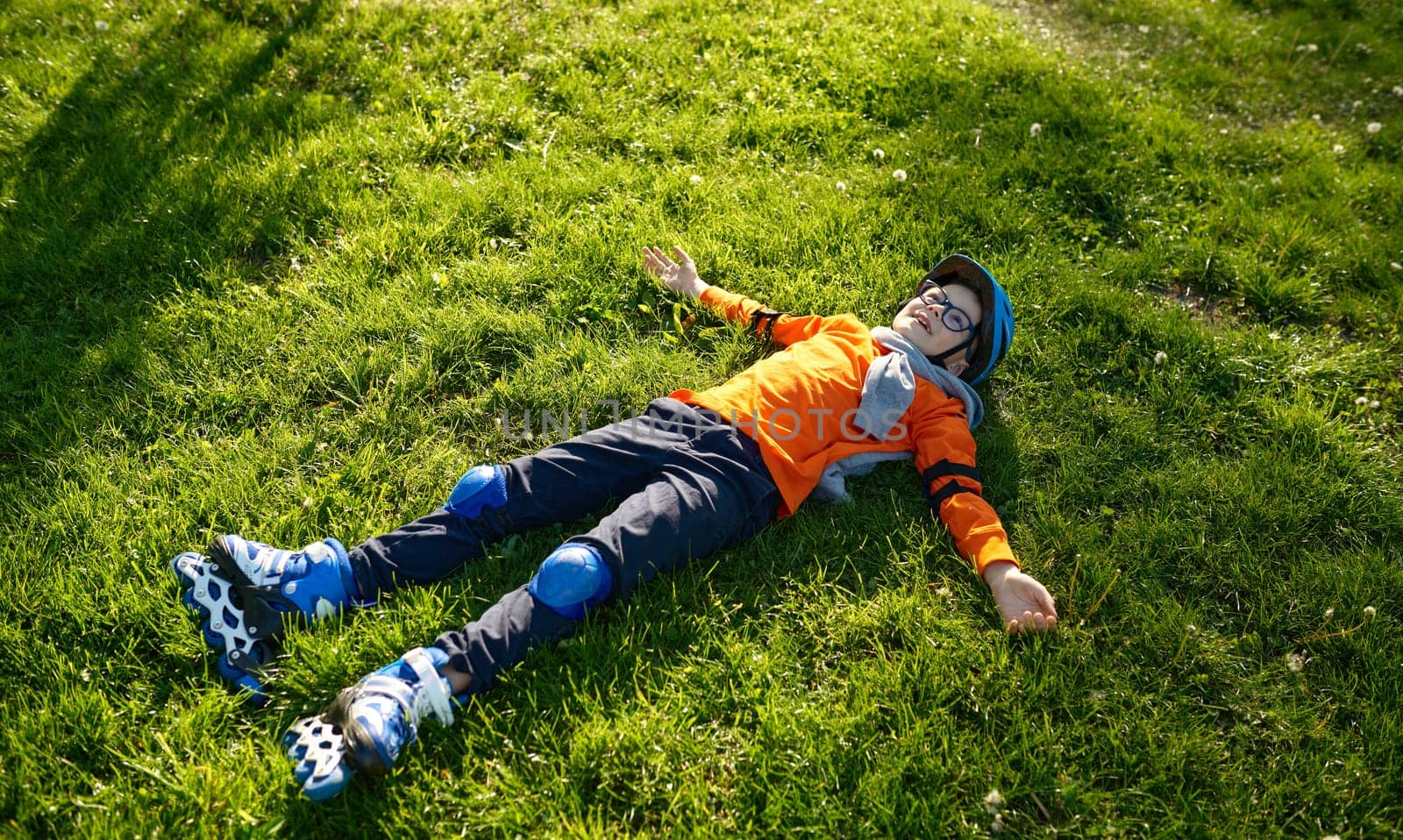 Happy tired little preschool boy wearing roller skates and helmet rest on green grass in urban park