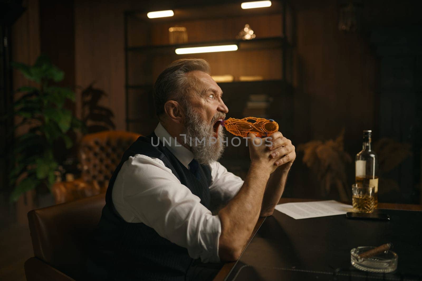 Funky elderly man trying to shoot himself with toy gun while sitting at desk table in home office. Business problems, bad contract, bankruptcy concept