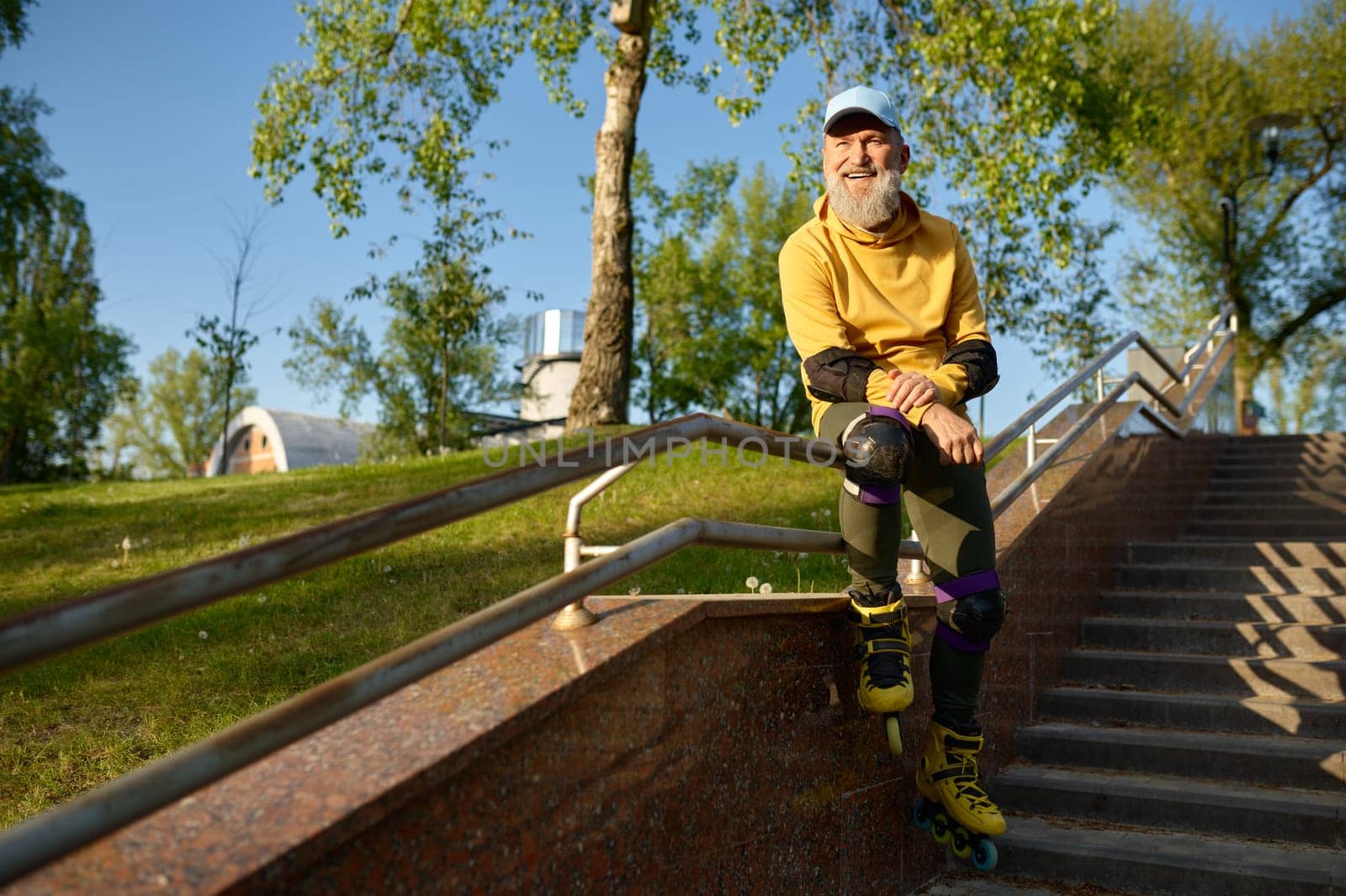 Happy smiling senior man wearing roller skates rest sitting on stair railing in urban park. Active time on retirement