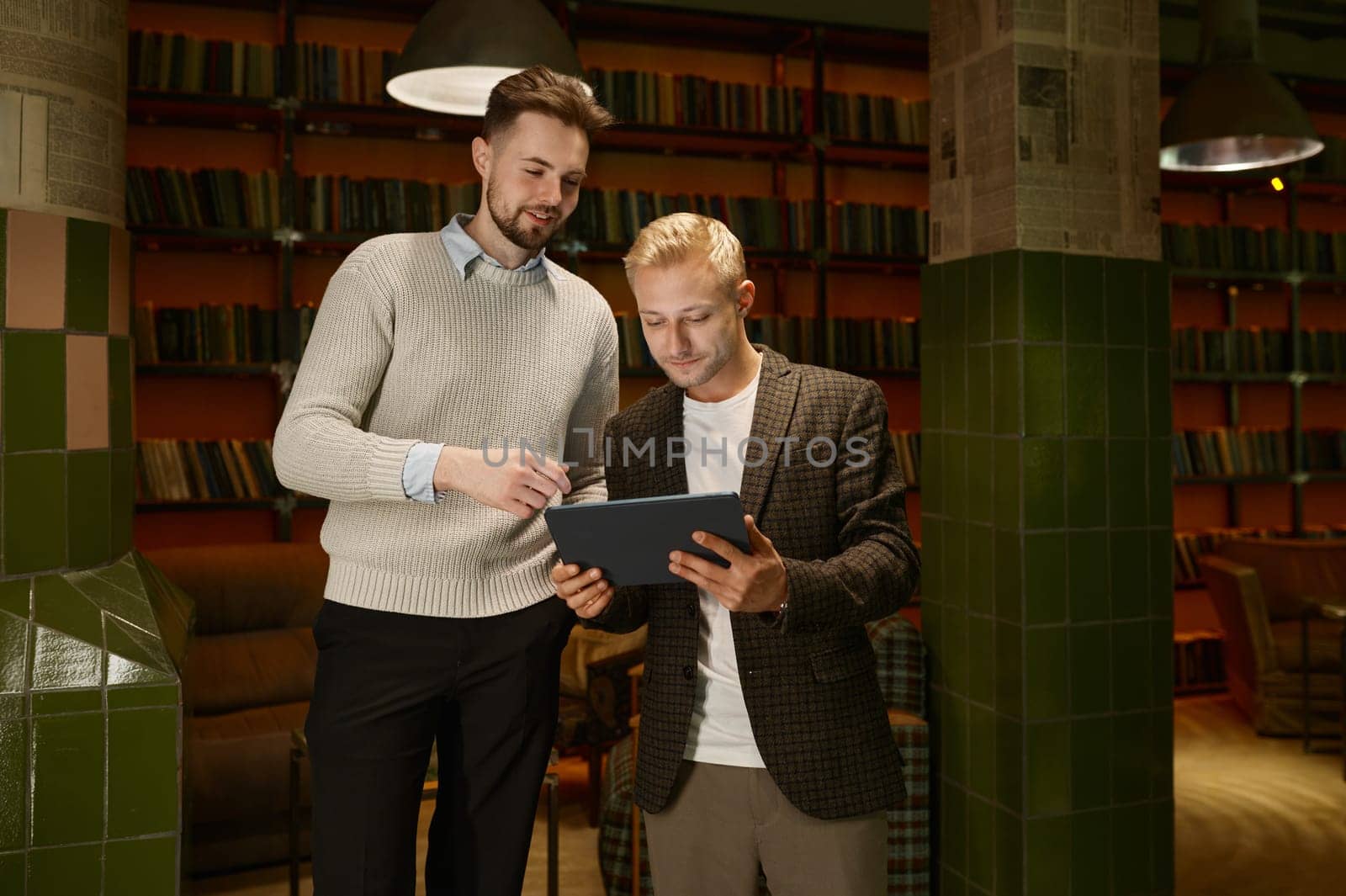 Two male business partners or web designers discussing project looking at mobile tablet while standing in office room of coworking space