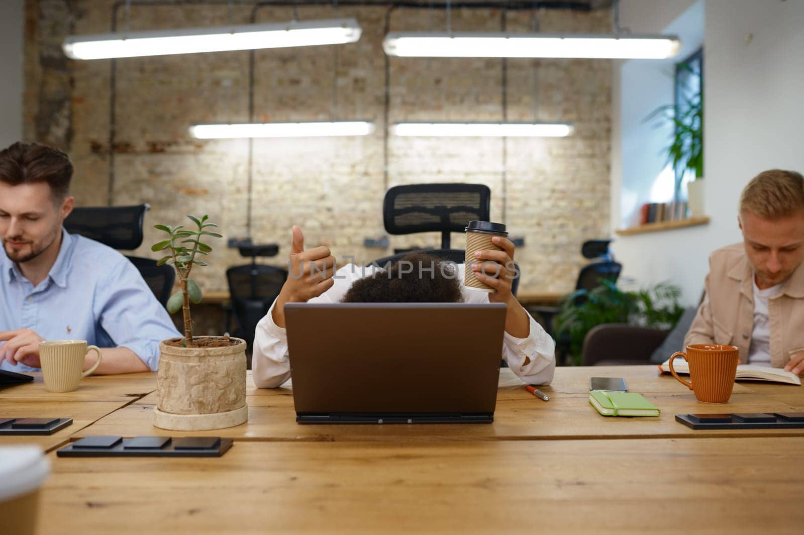 Woman office worker hiding head behind laptop gesturing thumbs up feeling tired and exhausted but trying to be positive and motivated. Coworking space with colleagues engaged in work