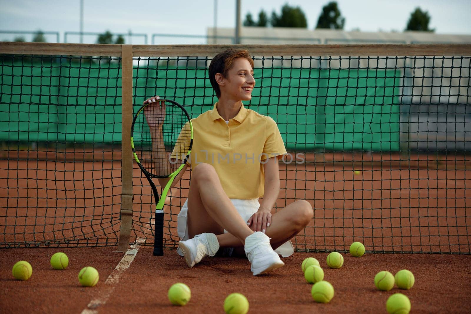 Attractive smiling woman tennis player with racket sitting on court among scattered balls. Cute woman relaxing after game training