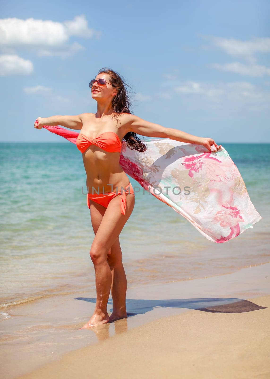 Young woman wearing orange red bikini and sunglasses standing on the beach waving silk scarf in wind behind her, sea background.