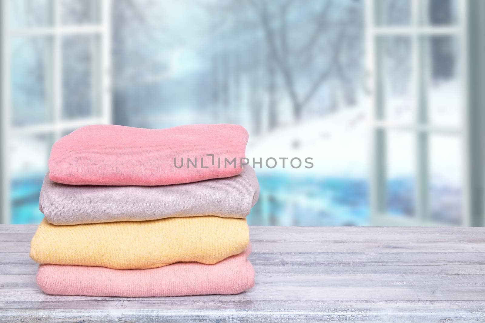Stack of winter clothes. A pile of colorful cozy warm sweaters on a wooden table against abstract blurred natural winter background. Autumn and spring clothes. Copy space.