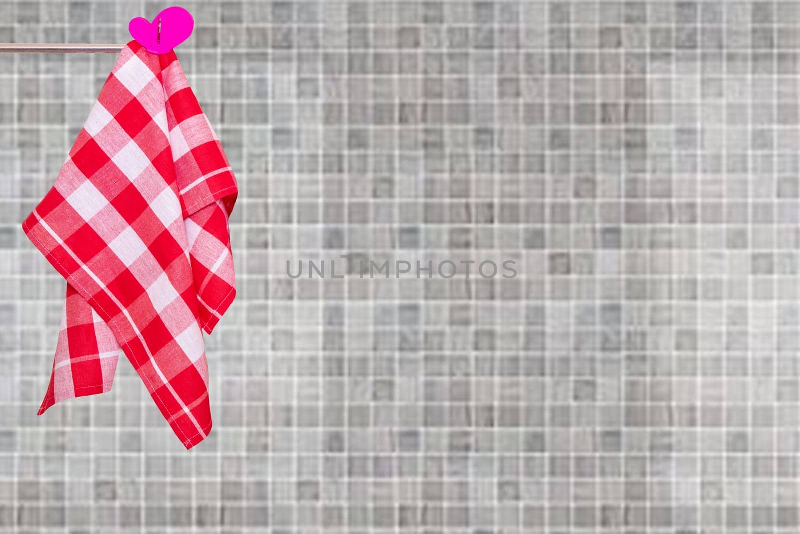 A red checked kitchen towel hanging on a clothes rail against abstract blurred beige gray mosaic ceramic tile wall. Space for your product display montage.