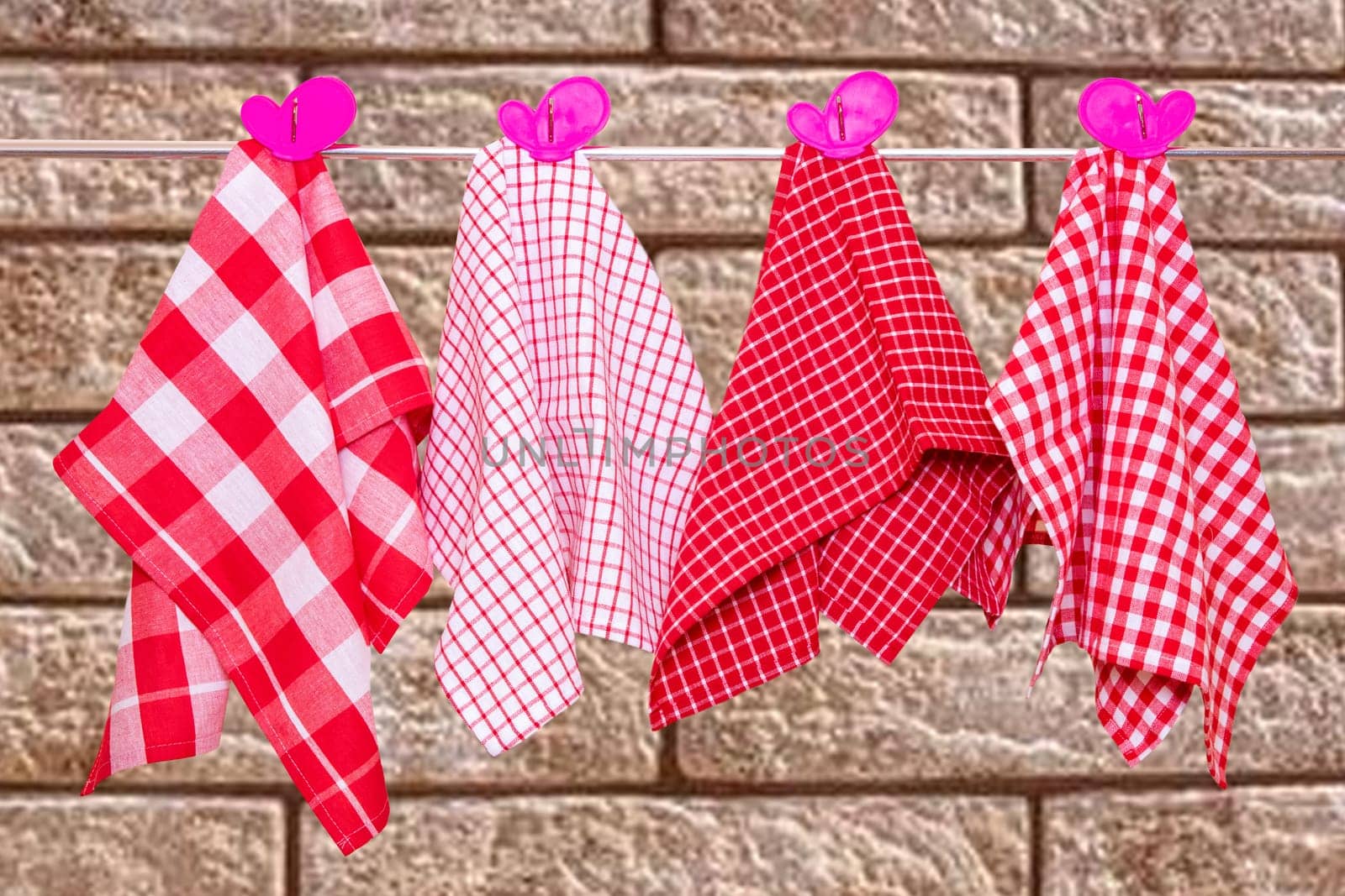 Hanging towels. Closeup of various red checkered kitchen towels with violet pink clothespins hang on a clothes rail against abstract blurred brick wall.