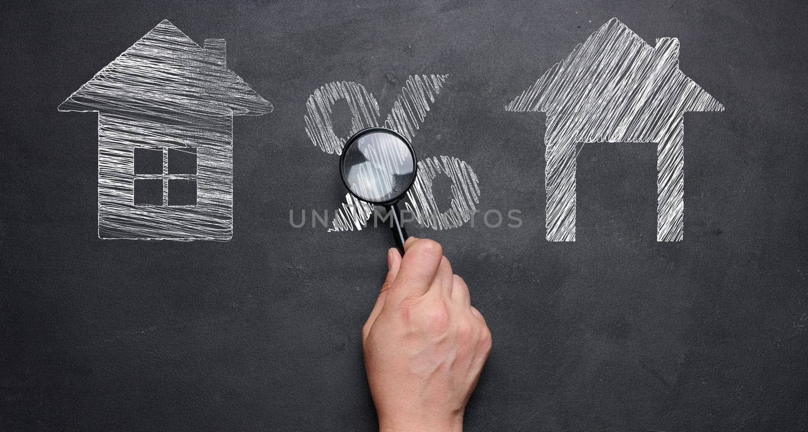 Chalk-drawn houses and a percentage sign on a blackboard, a female hand holding a magnifying glass. Concept of increasing or decreasing mortgage, rent.
