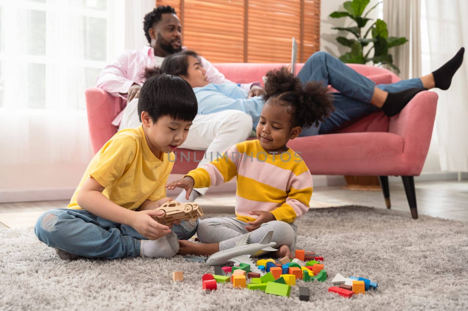 Cute kids playing with colored wooden blocks, airplane and cars on floor in living room while parents relaxing on couch with tablet. Happy family enjoying spending time together at homeEducational toys for preschool and kindergarten child and family entertainment concept.