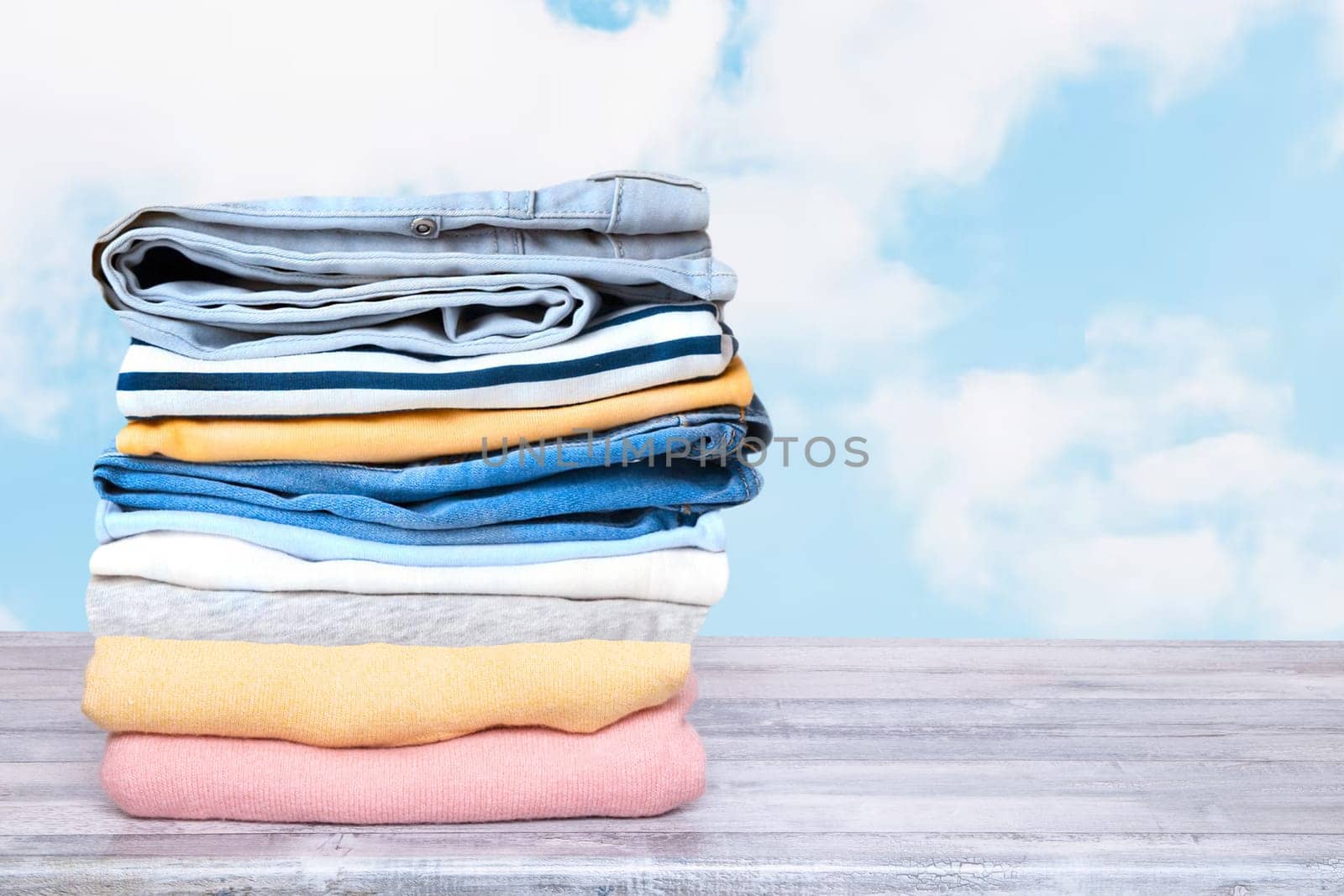 Stack of jeans. Closeup of a pile of colorful denim pants and colorful sweater on a bright table against abstract blurred natural light blue sky background. Space for your product display montage.