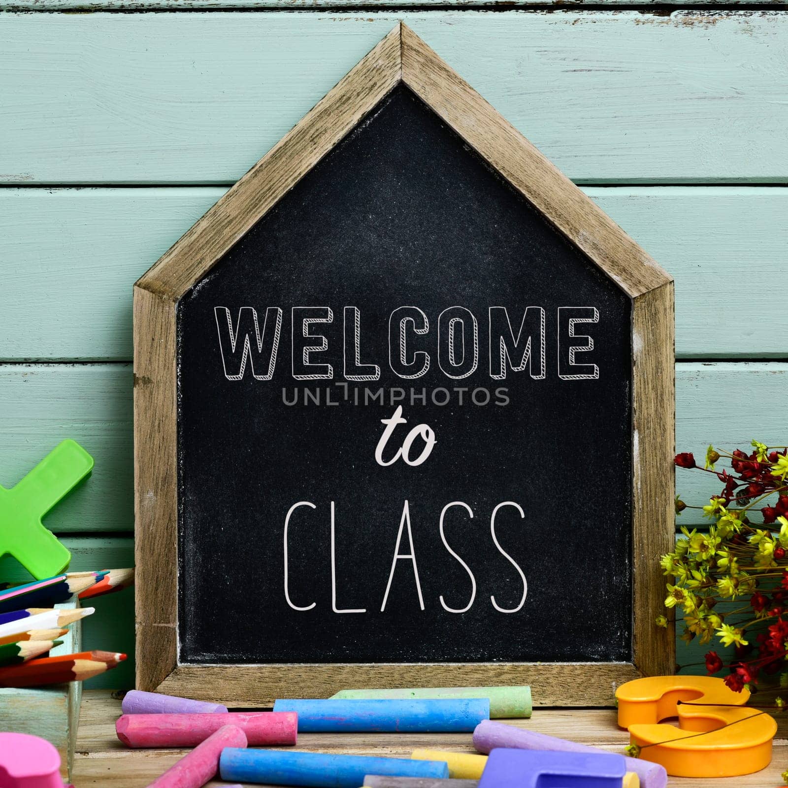 closeup of a house-shaped chalkboard with the text welcome to class written in it and a pile of pieces of chalk of different colors, against a pale green wooden background