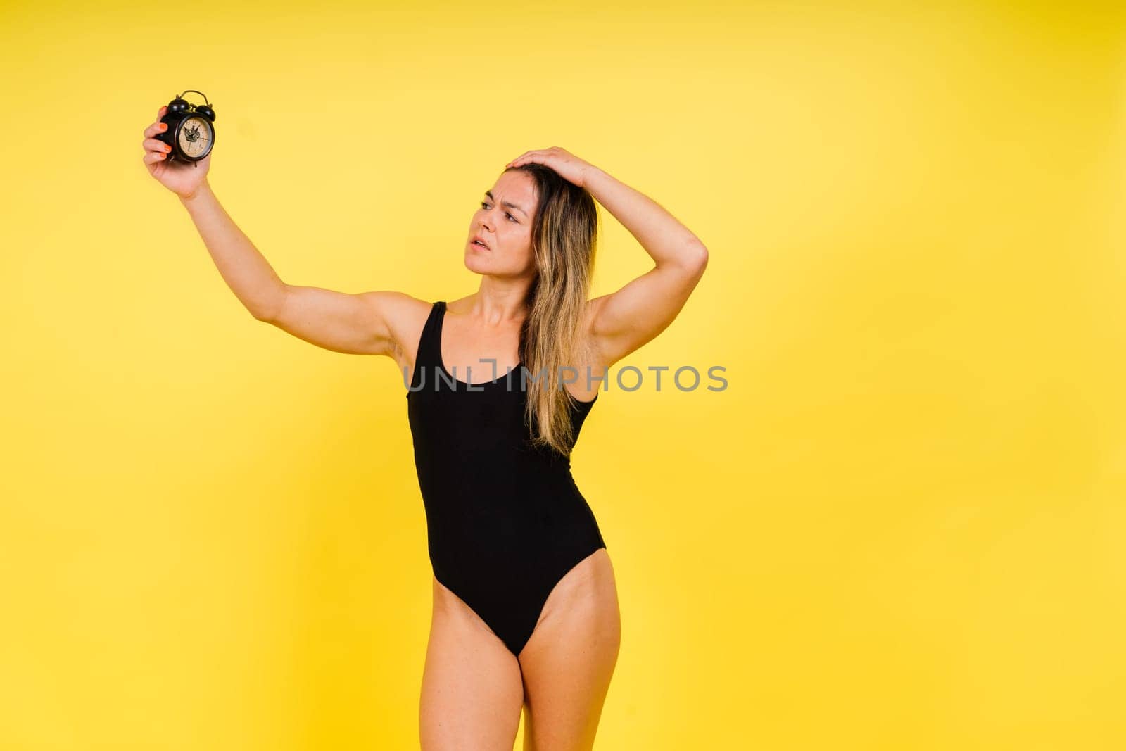 Female holding alarm clock, isolated on yellow. Young model in a black swimsuit.