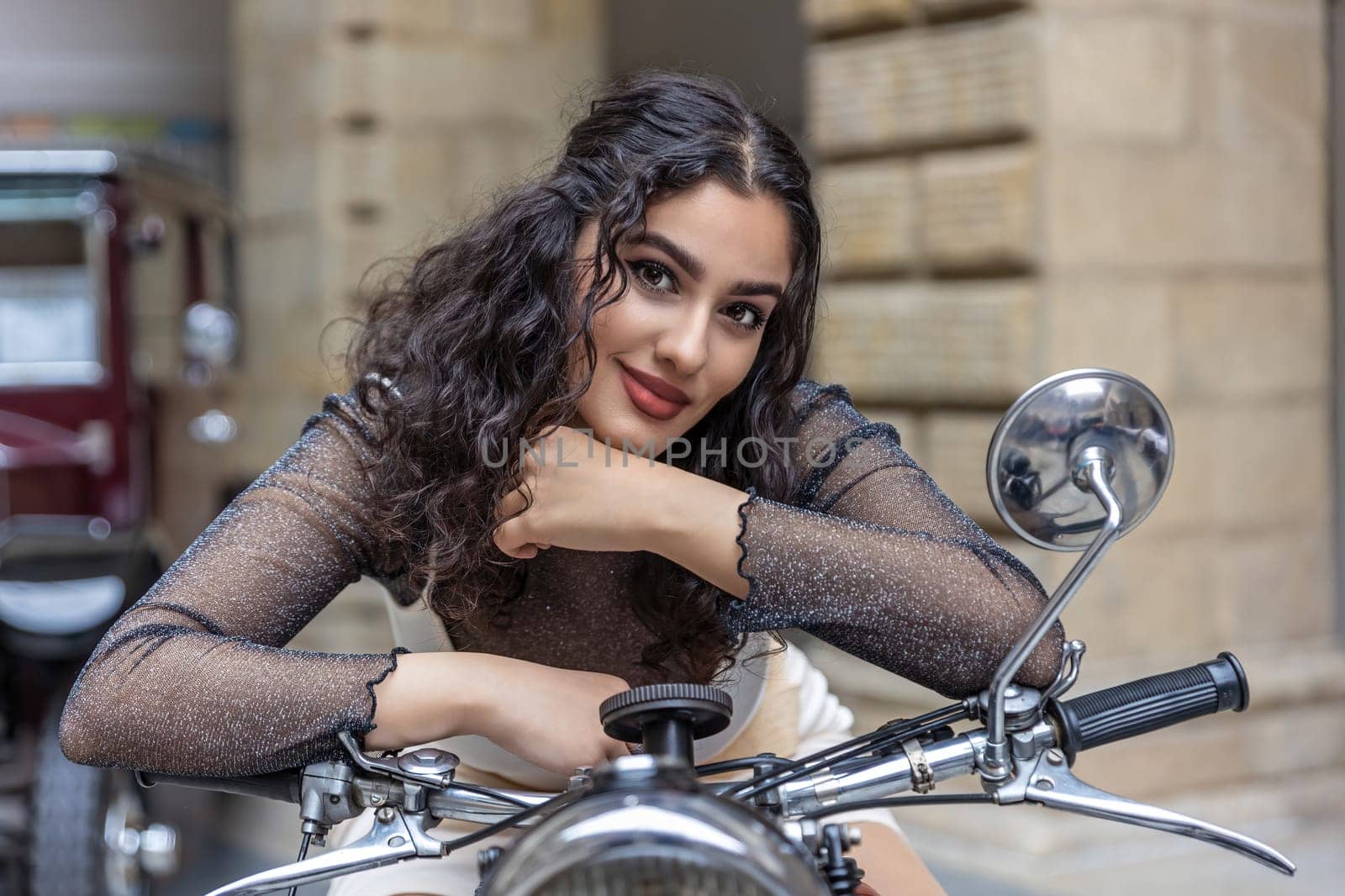 Front view of attractive tanned girl sitting on a vintage motorcycle looking at the camera leaning on the handlebars.  Horizontally.