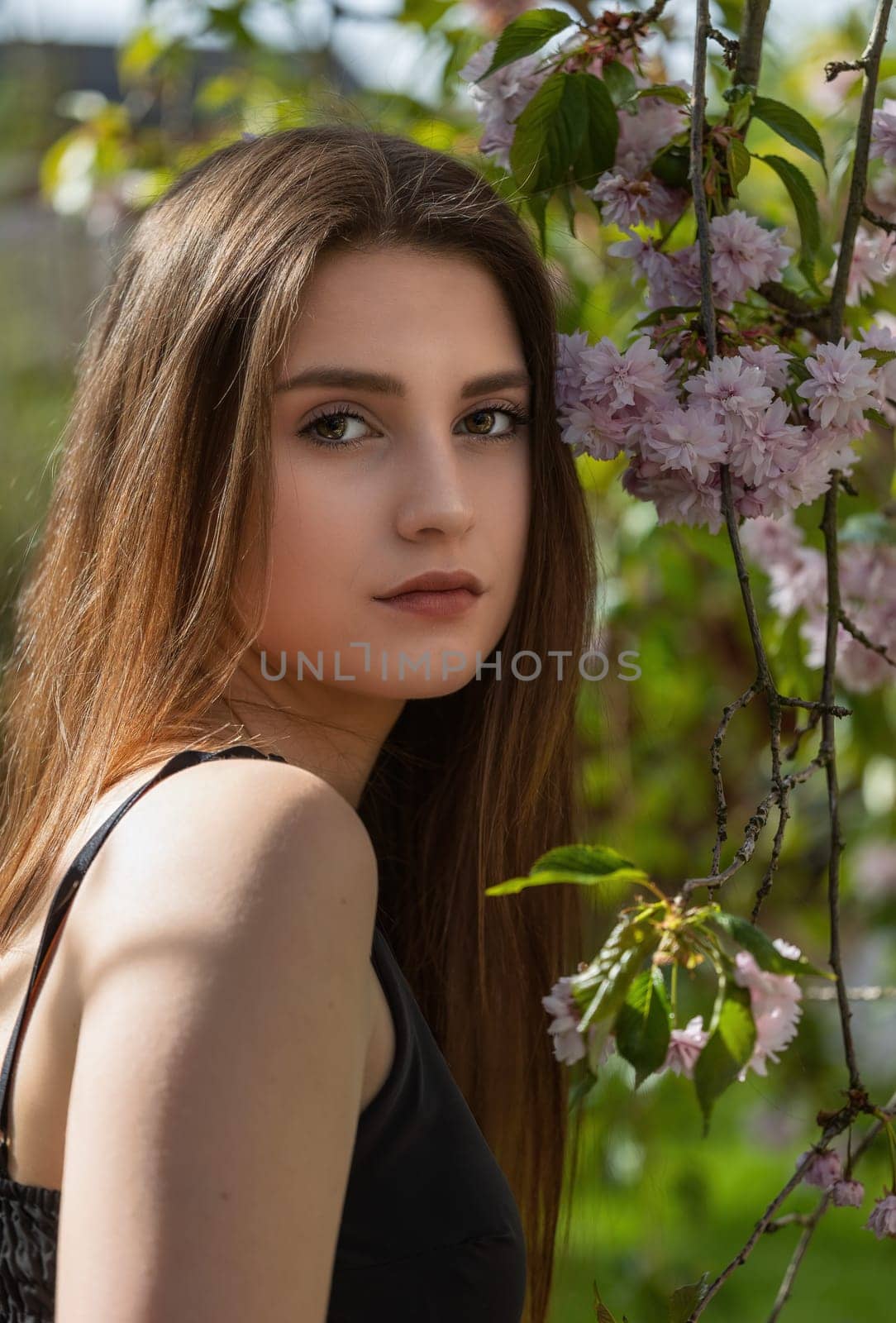 Cool brunette girl in black is posing on camera outdoors near sakura tree looking at the camera. Vertically.