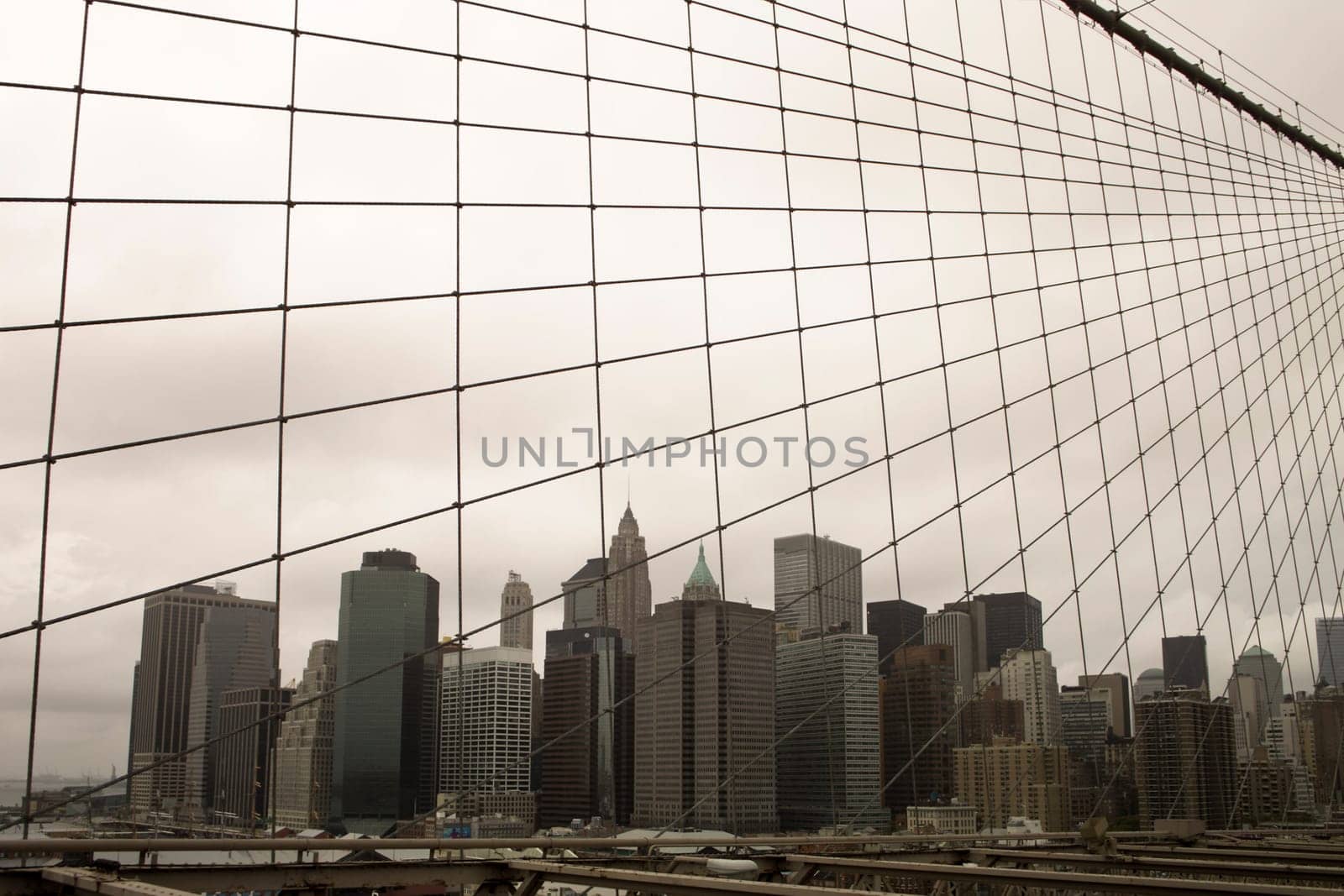A view of Manhattan downtown and the skyscrapers near Wall Street from the Brooklyn Bridge. The gloomy sky with leaden gray clouds is segmented by a web of cables from the suspension bridge.