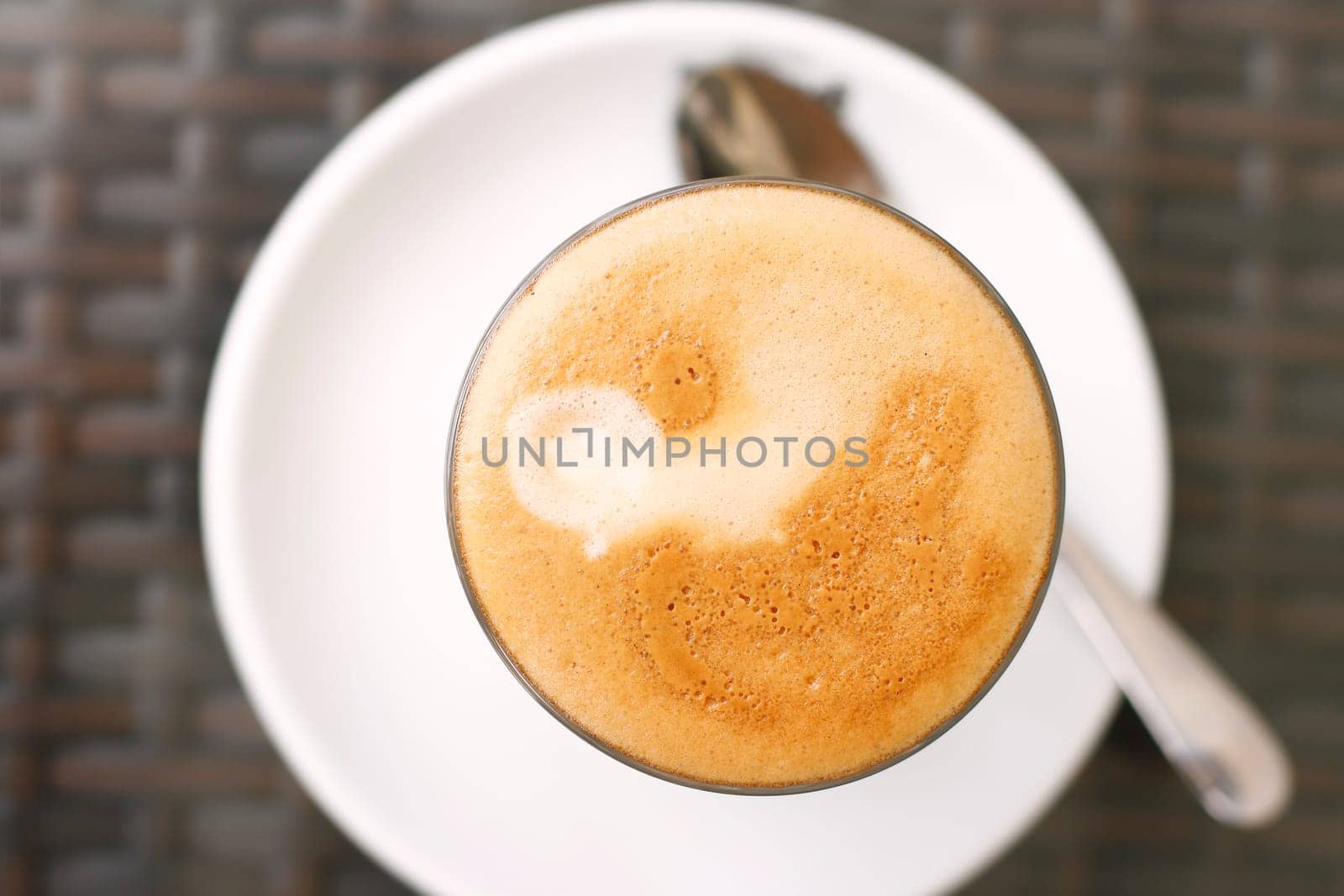A top down view of a latte with an in focus basic coffee art design in a glass framed by its out-of-focus white saucer and spoon.