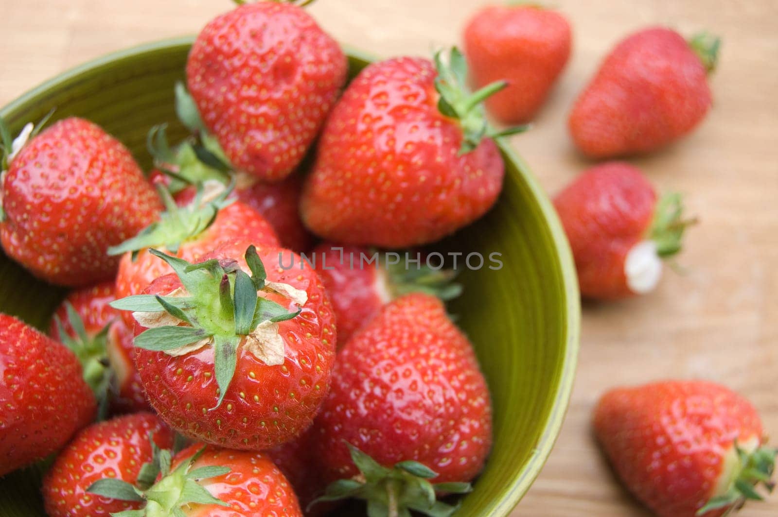 Overhead soft focus shot of red fresh strawberries in green bowl spilling over onto wooden board
