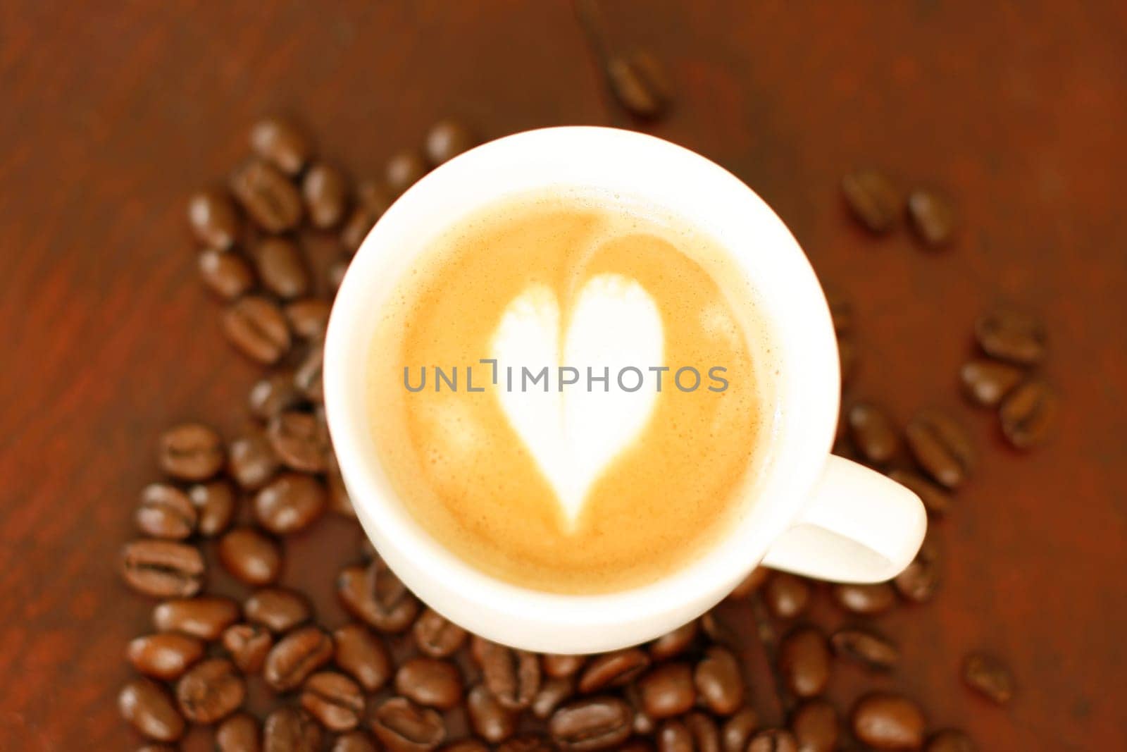 Top down view of a delicious piccolo latte in a small white cup with a heart shaped coffee art design sitting on a wooden table surrounded by roasted coffee beans.