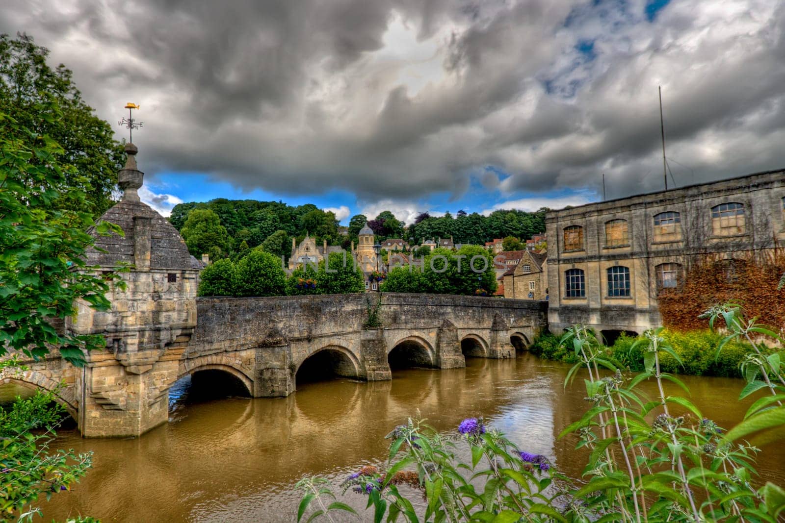 Bridge over the river Avon, Bradford-on-Avon, near Bath, England. The history of this bridge spans seven centuries. The "broad ford"" across the Avon, which gave the town its name, served as a crossing point until the original packhorse bridge was built in the 13th century. Two ribbed and pointed arches from this ancient bridge survive on the eastern side of the river. Various repairs and alterations have been carried out over the centuries, including the widening of the bridge to twice its original width. John Aubrey visited the town in 1660 and commented, ""Here is a strong and handsome bridge in the middest of which is a little chapell for masse as at Bathe."" This chapel on the bridge dates from the 17th century and was converted into a prison or ""Blind House"" at a later date."