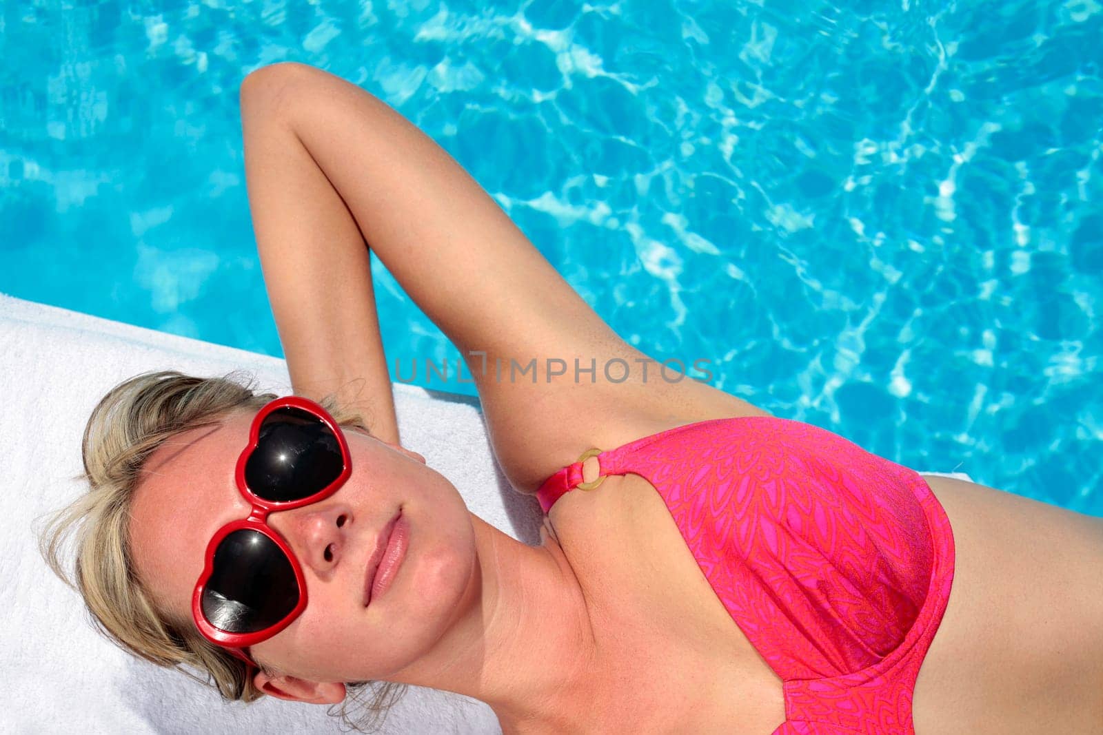 Woman in heart-shaped sunglasses besides a bright blue swimming pool
