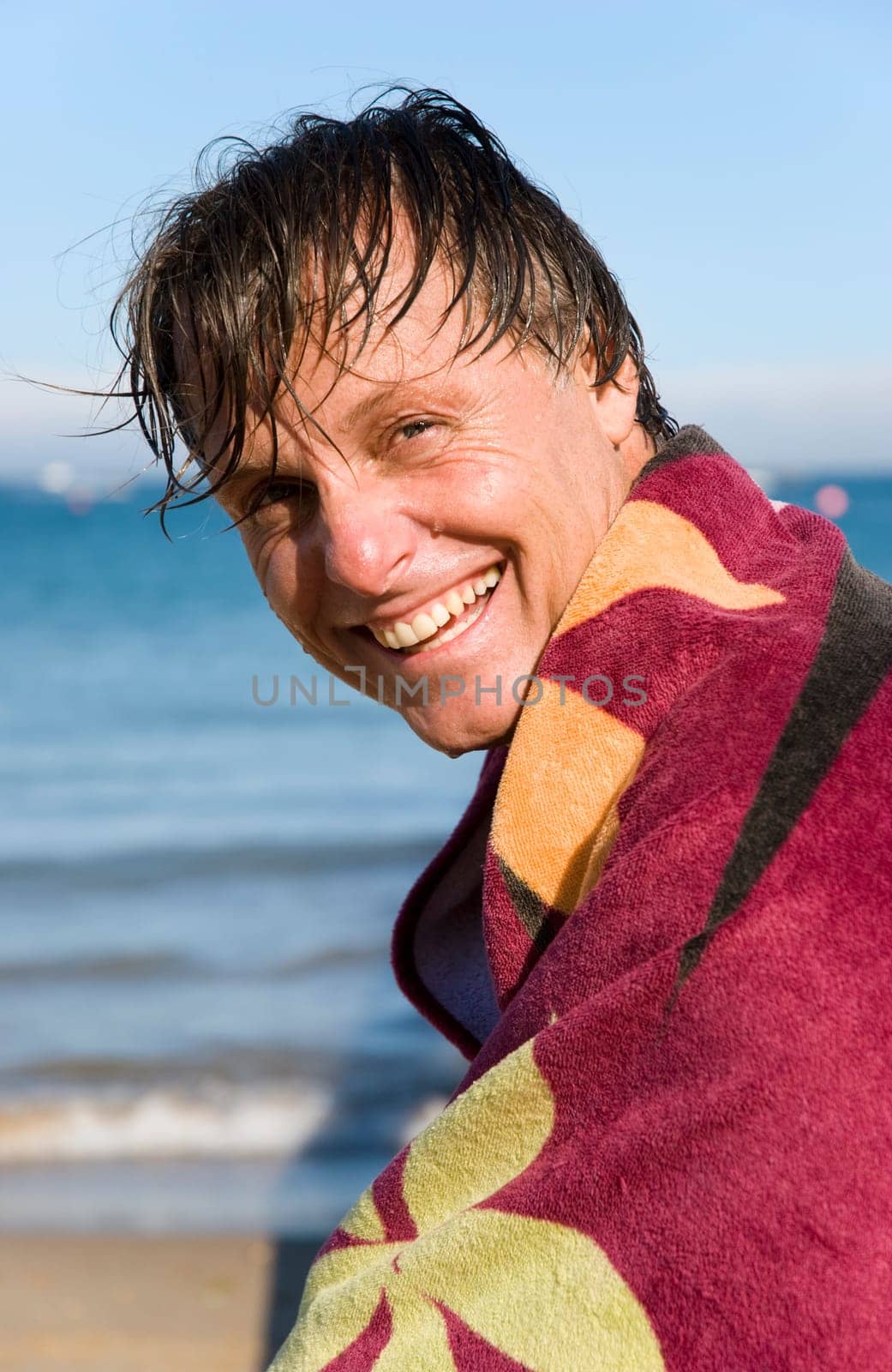 A handsome smiling forties man is sat on a beach with a towel wrapped around him to keep warm.