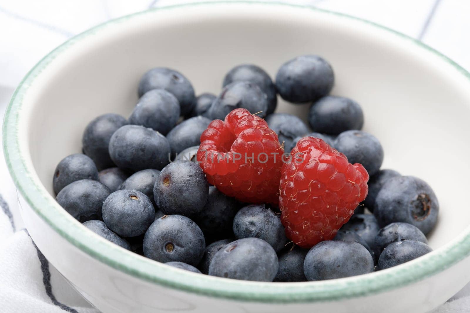 A close-up of fresh blueberries and raspberries in a bowl sitting on a white kitchen cloth.