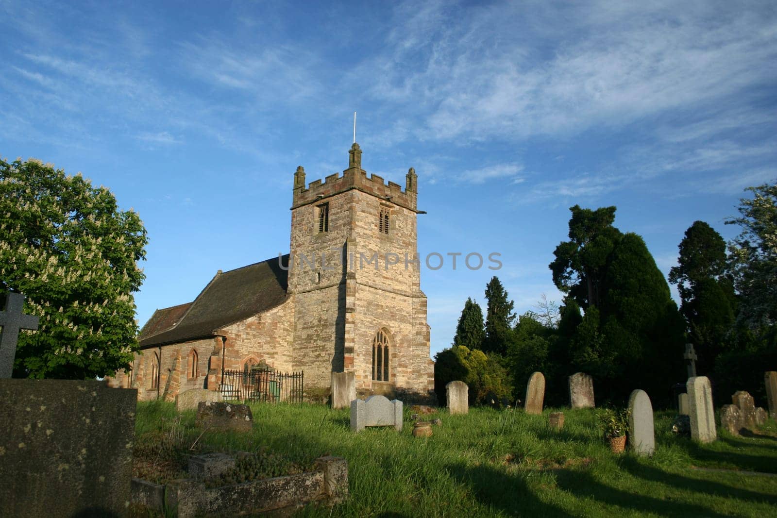 The church of St John the Baptist in the village of Feckenham in Worcestershire, England. Late afternoon sun casts long shadows from the grave stones and lights the church against the blue sky.