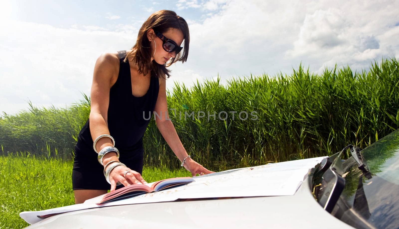Young woman, studying a map and a guide book on the hood of her car on a bright summer day