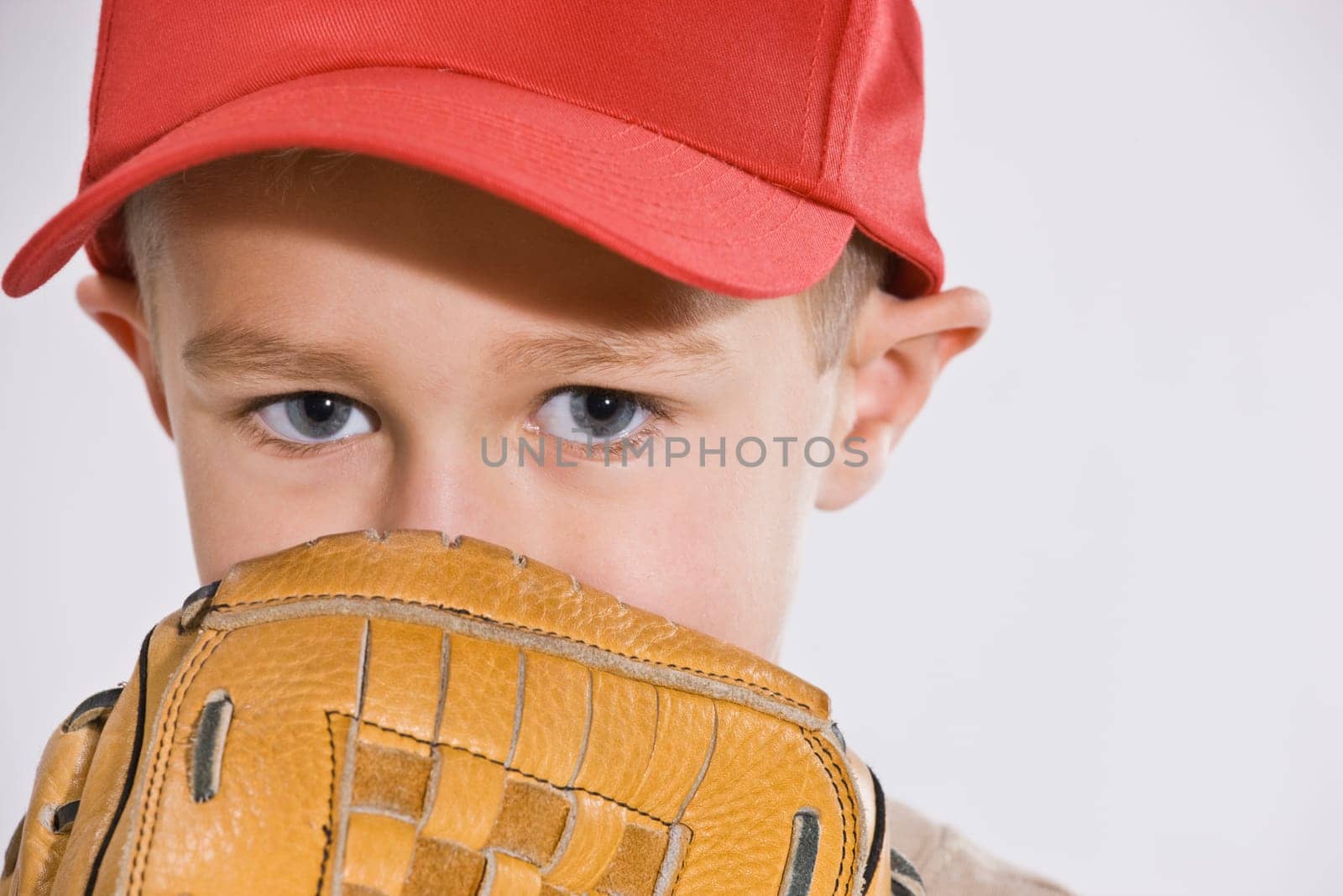 A young boy is wearing a baseball cap and holding a baseball mitt up to his face.  Horizontally framed shot.